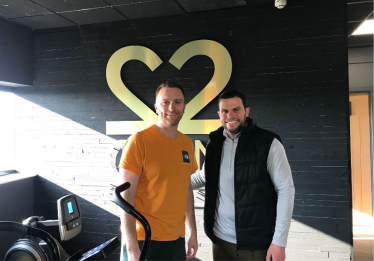 Matt and owner Garry smile in front of the Gyms logo, mounted to the wall behind them.