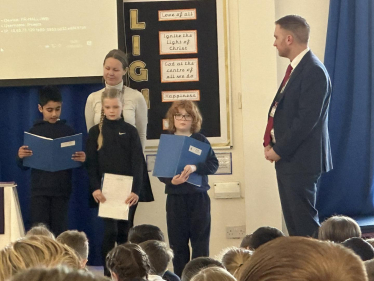 Matt Vickers MP stands on stage with school children who are reading aloud.